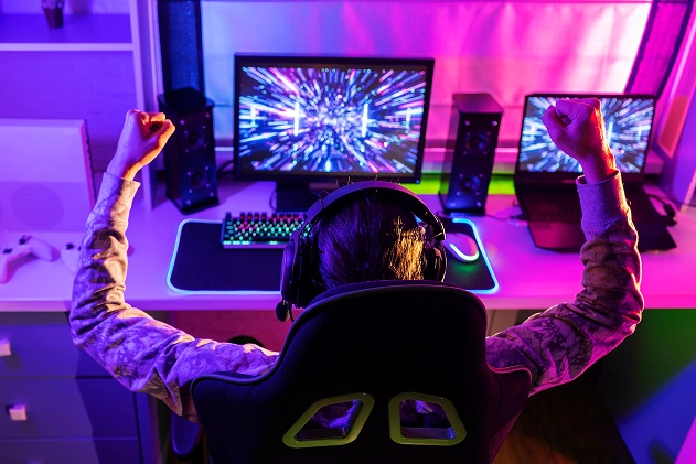 Um jogador, com os braços erguidos, celebra uma vitória em frente a um monitor de computador numa sala bem iluminada.